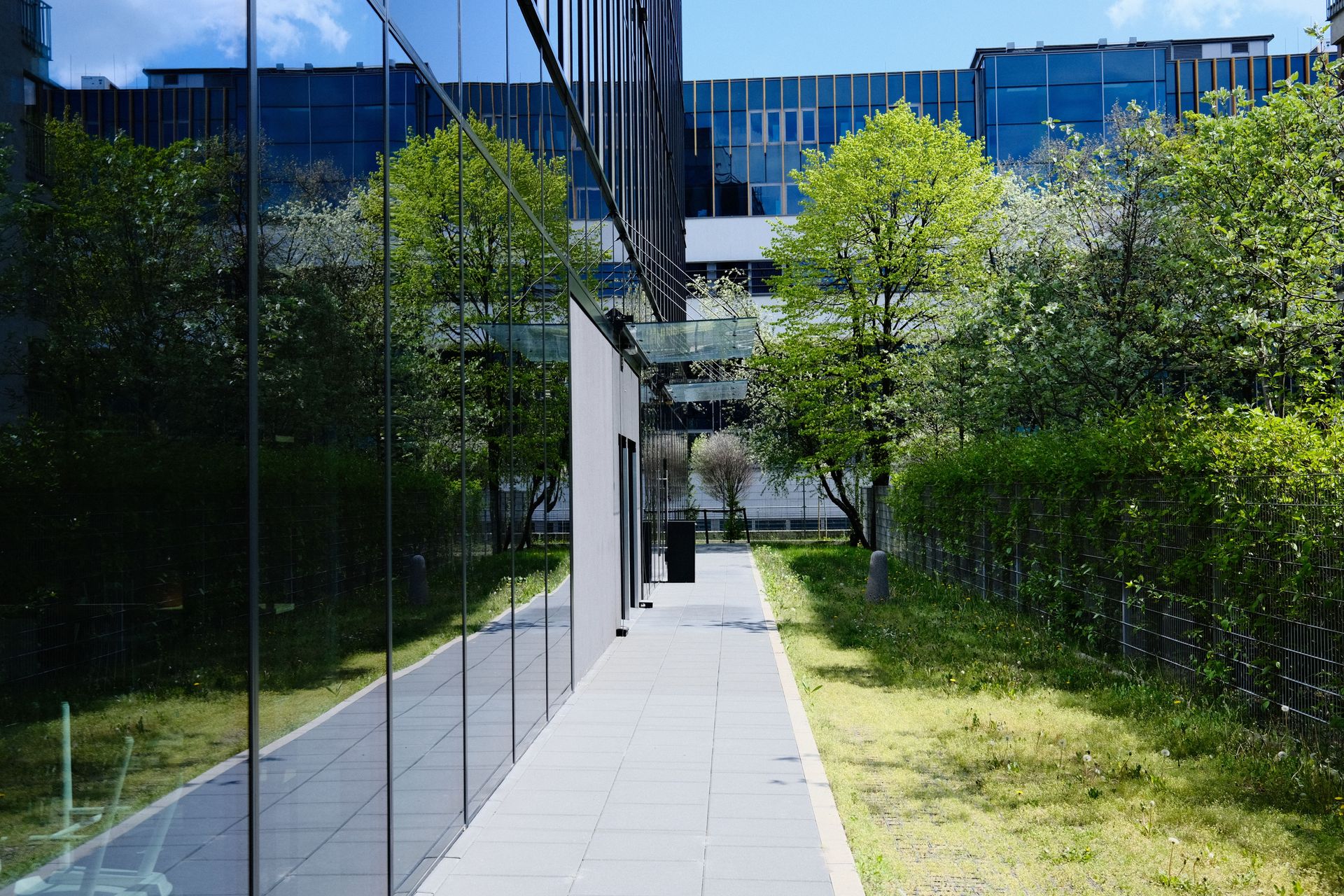 Modern glass building with pedestrian walkway and greenery, reflecting urban architecture.